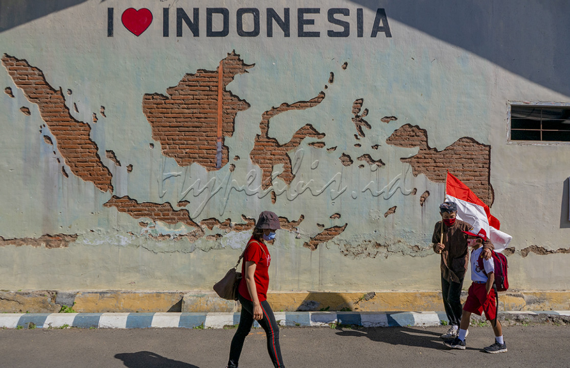 Seorang Anak SD dan Kakeknya yang bermasker membawa bendera Merah Putih berjalan menuju sekolah melintasi tembok bergambar peta Indonesia