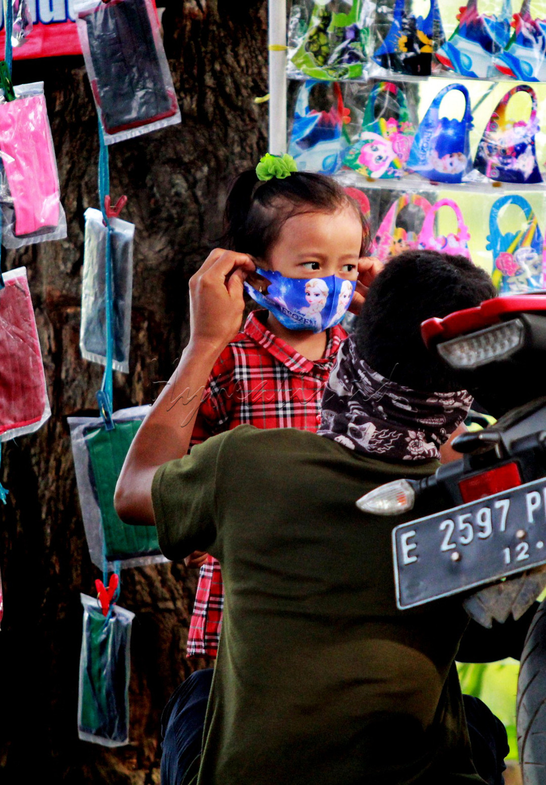 Seorang bapak tengah memakaikan masker kepada anaknya, usai membeli masker di tepi jalan. Ini dilakukan demi melindungi anaknya agar tetap aman dan merdeka dari Covid-19