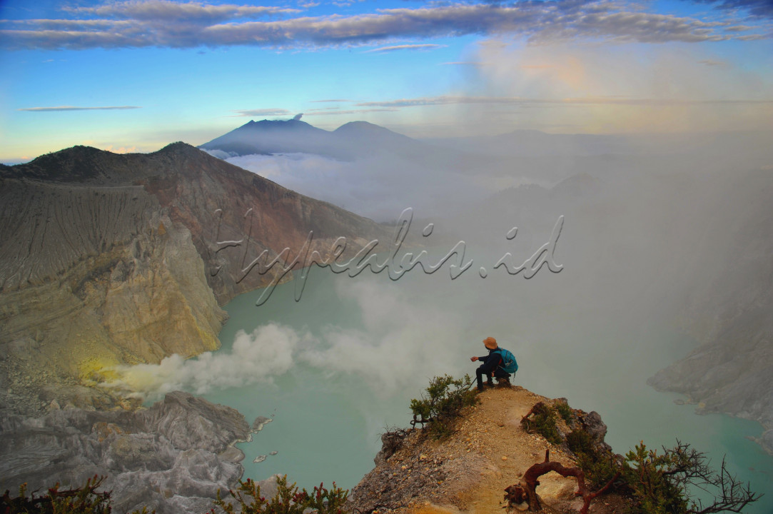 Seorang pendaki gunung sedang menikmati pesona Kawah Ijen saat menjelang matahari terbit. Kawah Ijen dari atas puncak Gunung Ijen terlihat sangat indah dengan danau besar berwarna hijau kebiruan serta kabut dan asap belerang yang sangat memesona. Gunung Kawah Ijen atau Kawah Ijen terletak di antara Kabupaten Bondowoso dan Kabupaten Banyuwangi yang merupakan salah satu gunung aktif di Jawa Timur. 